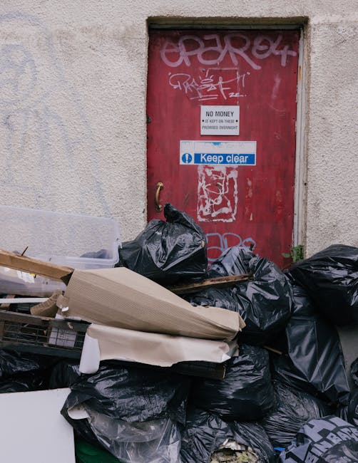 A pile of discarded household items and debris located on an outdoor paved area adjacent to a brick wall. The heap includes wooden furniture such as a dismantled, weathered wooden bed frame or slats p
